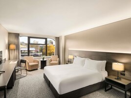 Habitación de hotel con una cama grande, dos sillones, un escritorio y vistas a la ciudad.