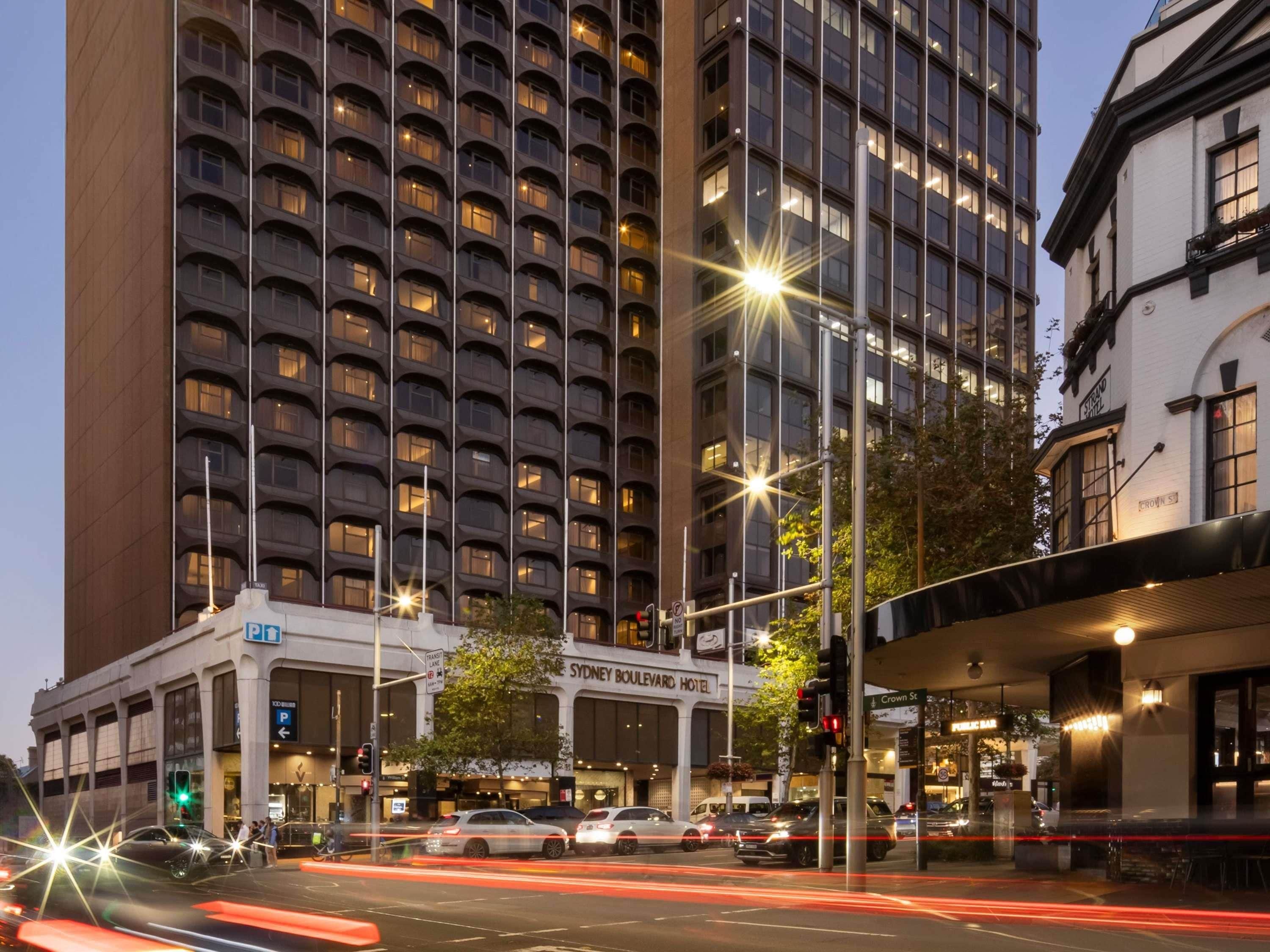 A multi-story hotel with a modern facade, a prominent sign for the Sydney Boxvillard Hotel, and a street with vehicles and traffic lights.