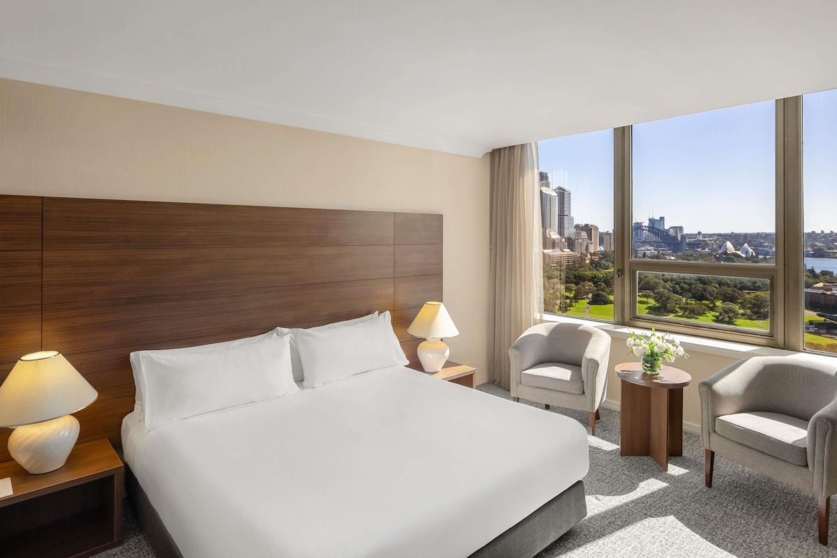 A hotel room with a large bed, two bedside tables with lamps, a chair, and a view of a cityscape.