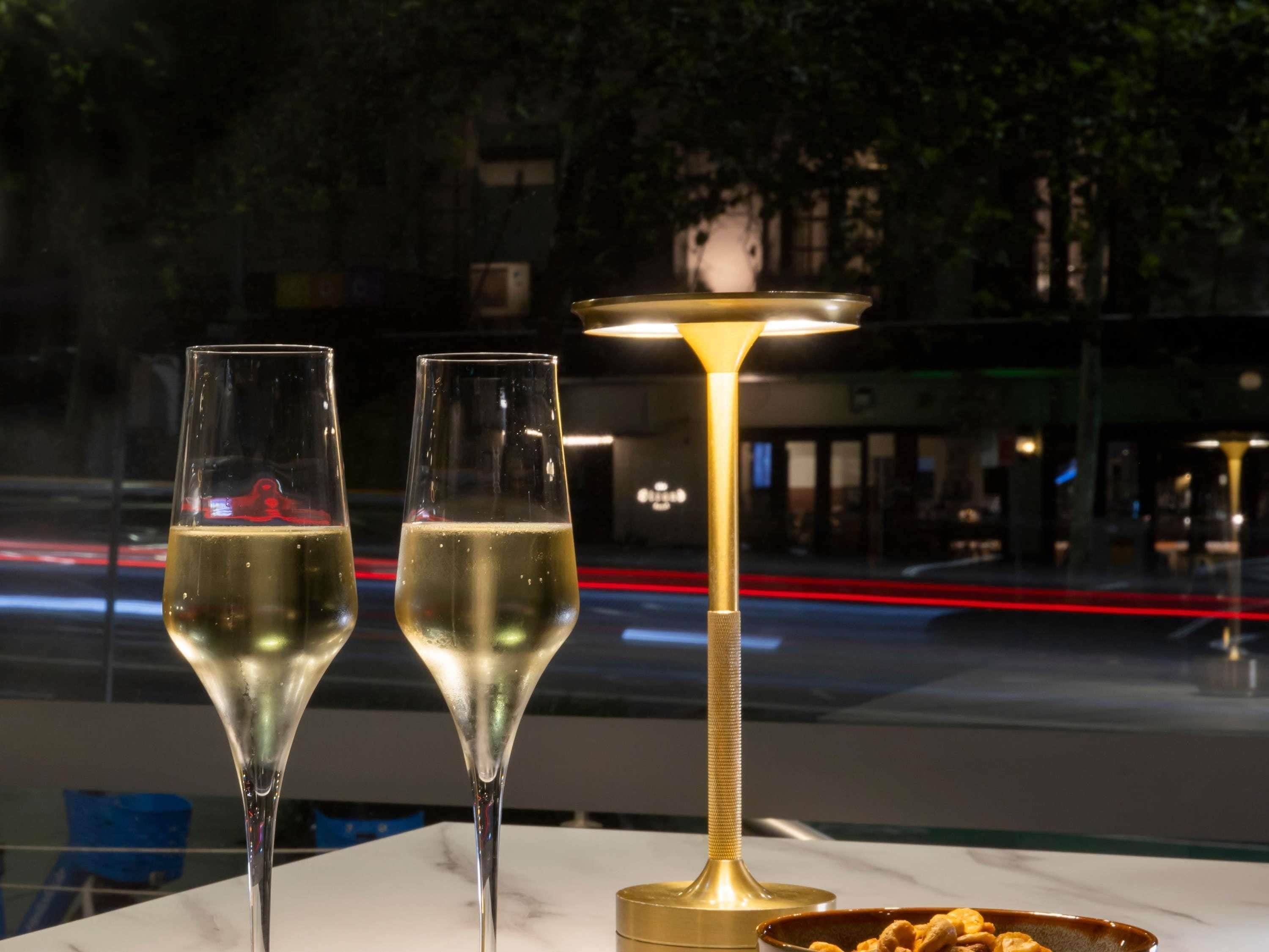 Två glas champagne på ett bord med en guldfärgad lampa, en skål med snacks och utsikt över en stadsgata i natt.