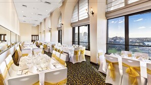 A banquet hall with round tables, white chairs with gold sashes, and large windows with a city view. - The Sydney Boulevard Hotel by Accor (Woolloomooloo)