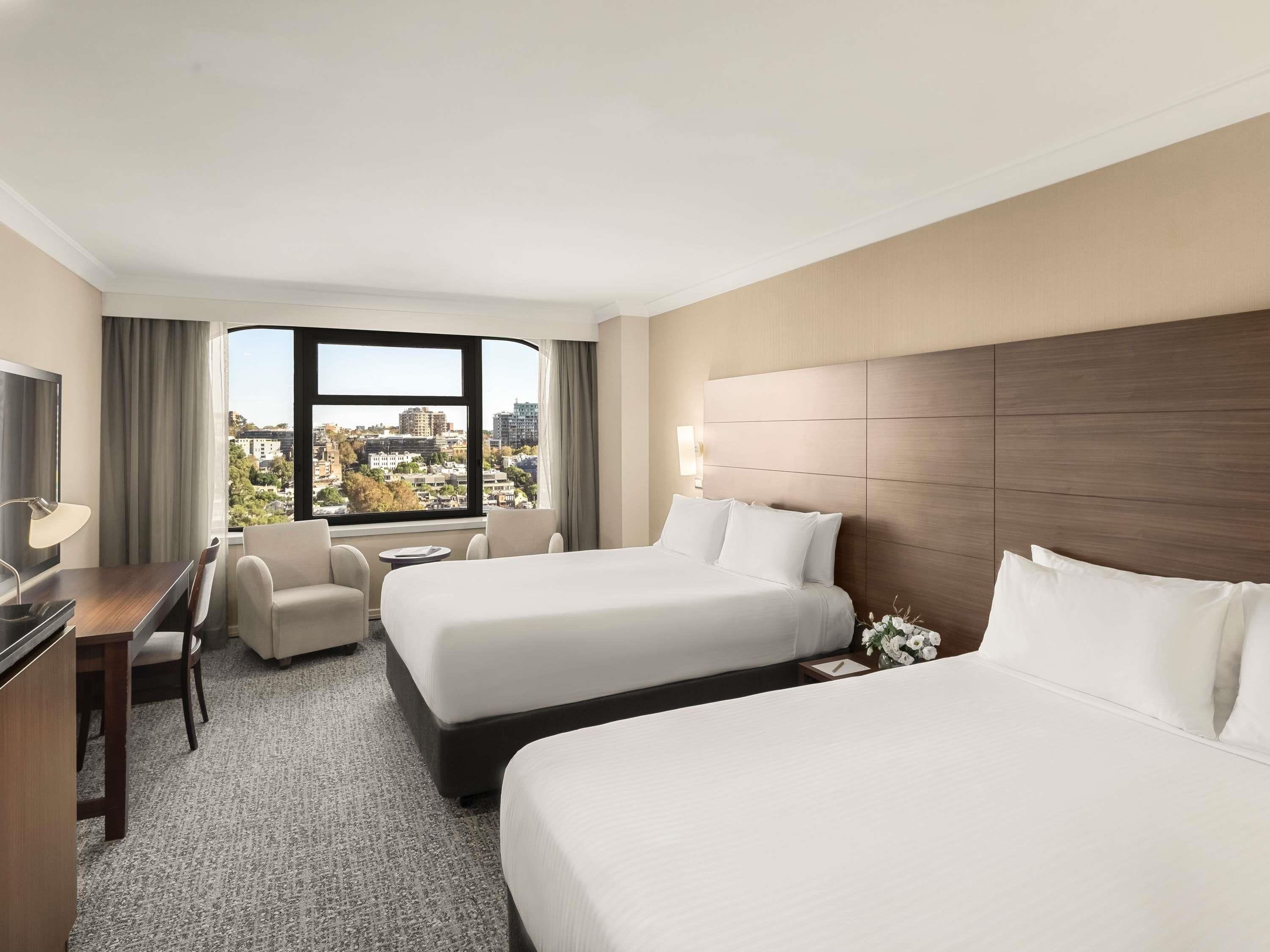 Une chambre d’hôtel avec deux lits, un bureau, une chaise, une petite table et une grande fenêtre donnant sur la ville.