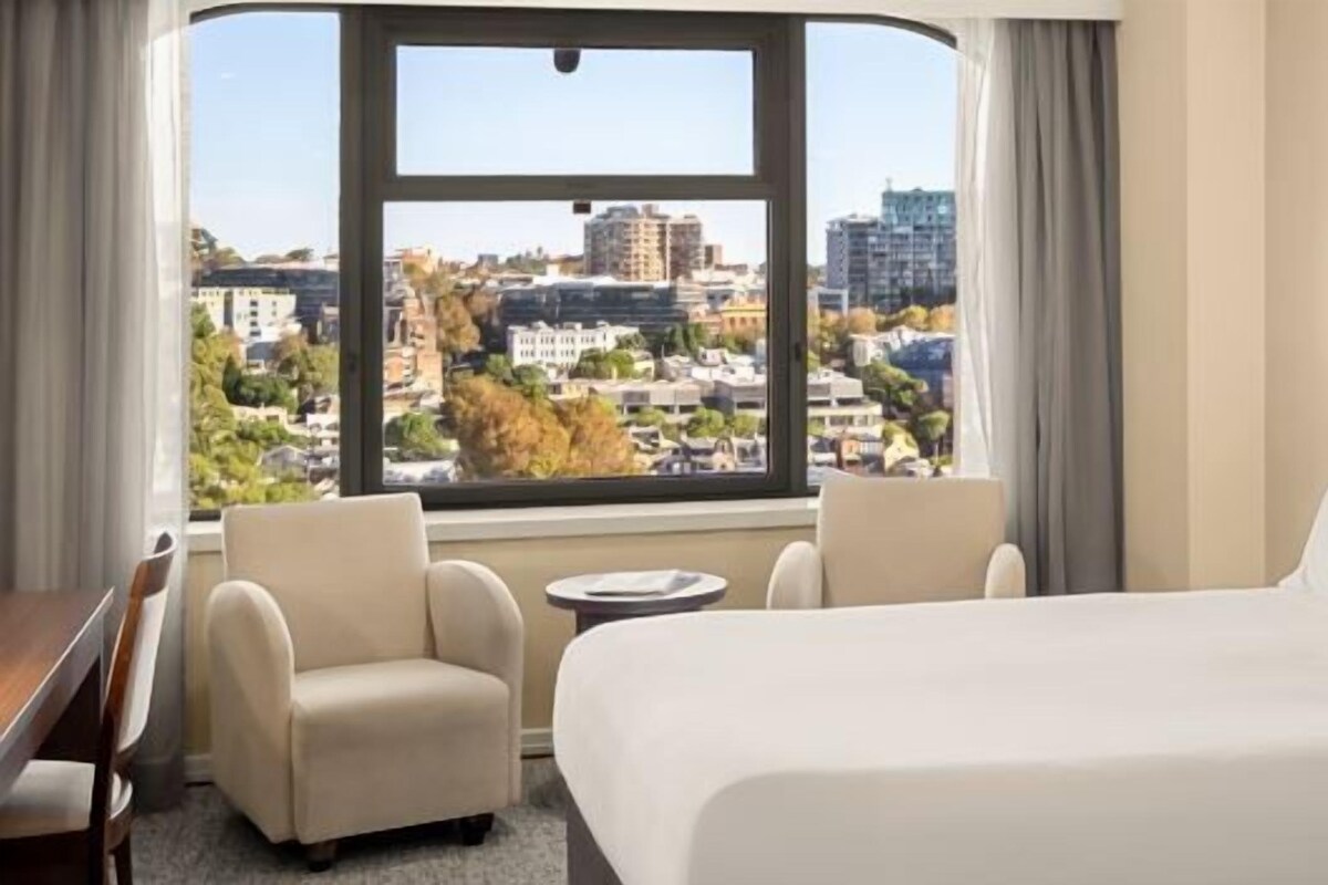 A hotel room with a large bed, a desk, two chairs, and a view of the city through a large window.