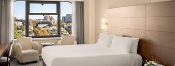 A hotel room with a large bed, a desk, two chairs, and a view of the city through a large window.