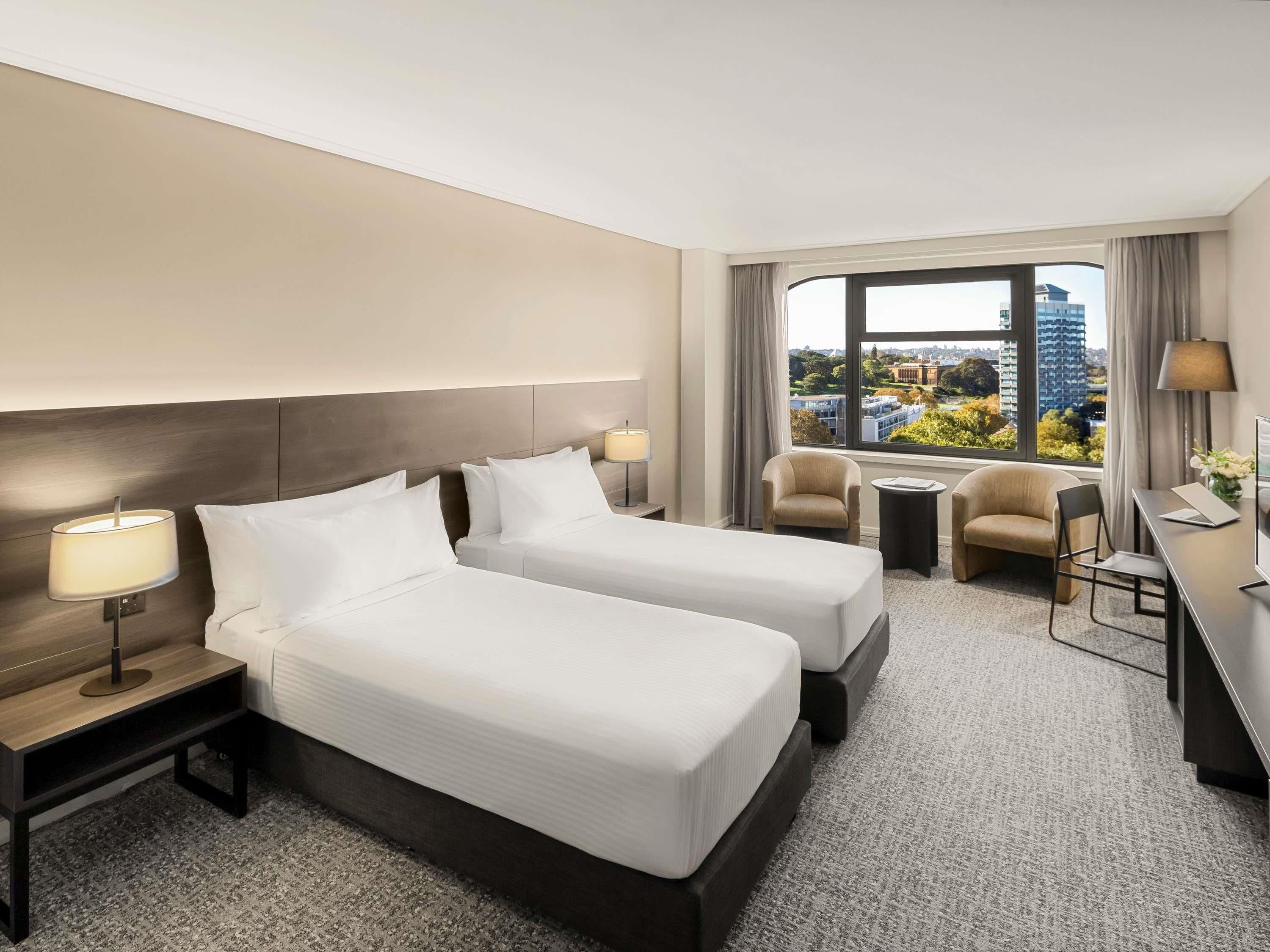 Habitación de hotel con dos camas, un escritorio, una silla, una lámpara y vistas a la ciudad.