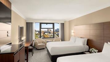 Habitación de hotel con dos camas, un escritorio, una silla, un televisor y un ventanal con vistas a la ciudad.