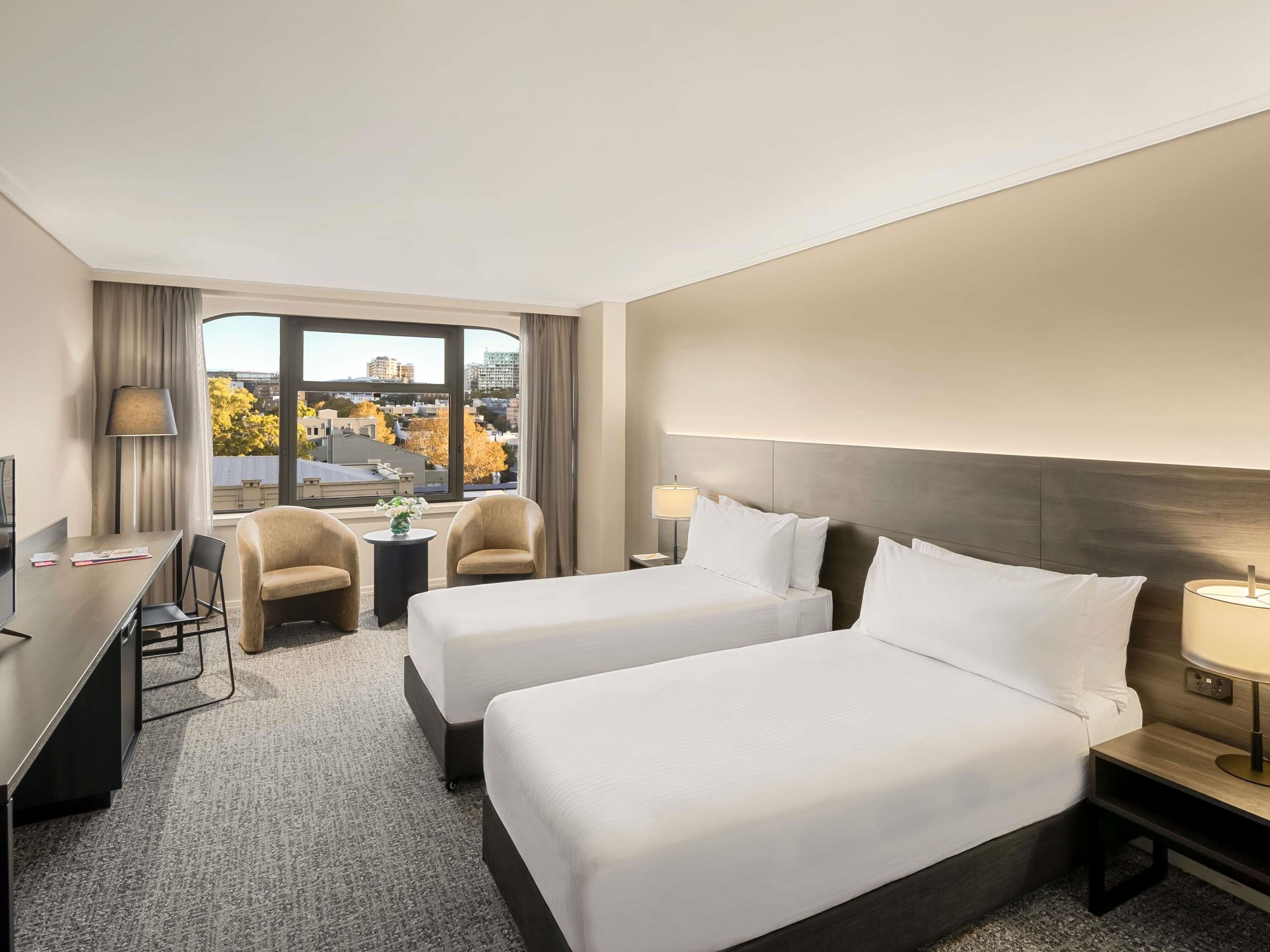 A hotel room with two beds, a desk, a chair, a lamp, and a view of the cityscape.