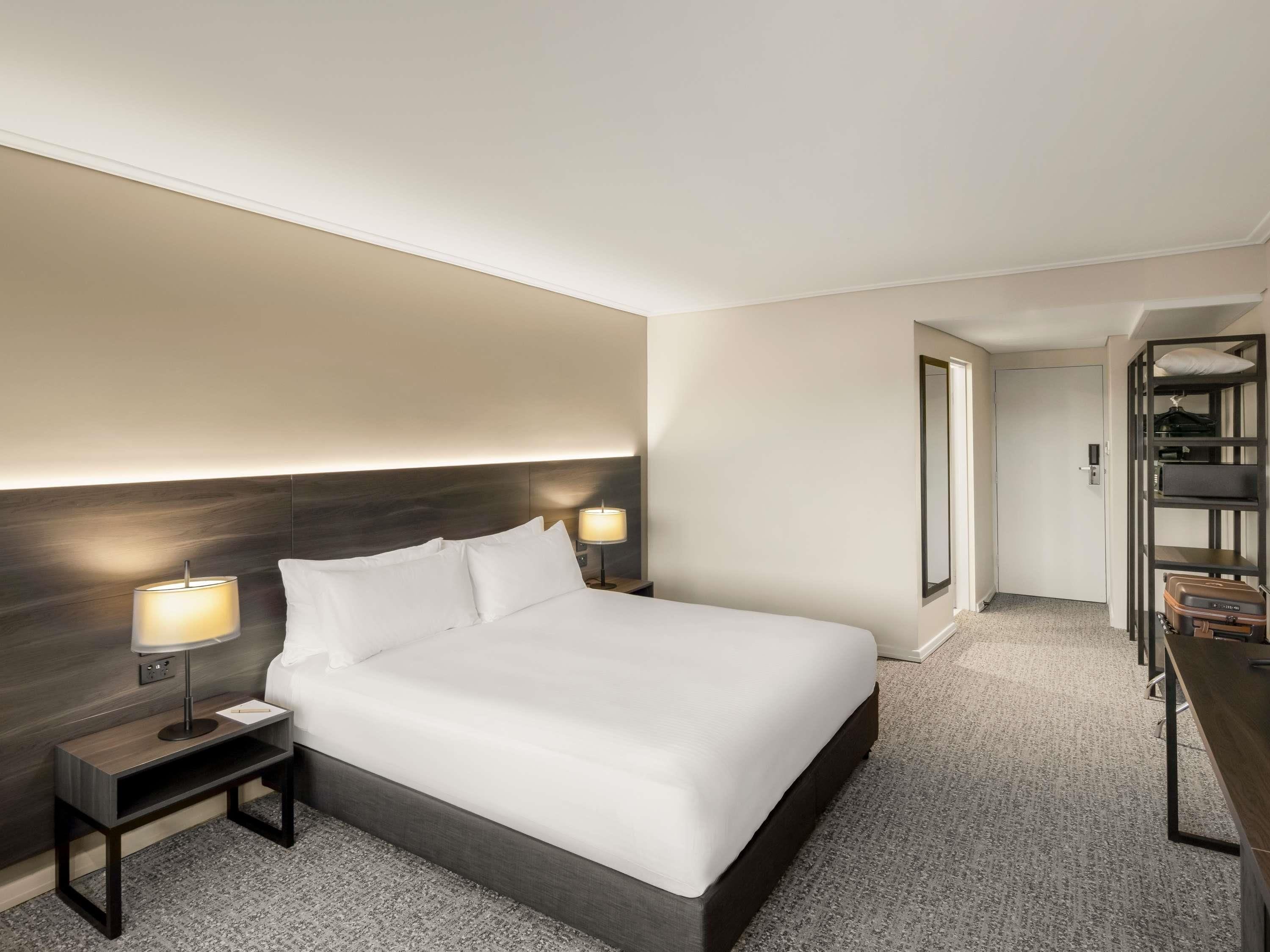 Una habitación de hotel moderna con una cama grande, dos mesitas de noche con lámparas, un cabecero oscuro, un armario y una puerta que da a otra habitación.