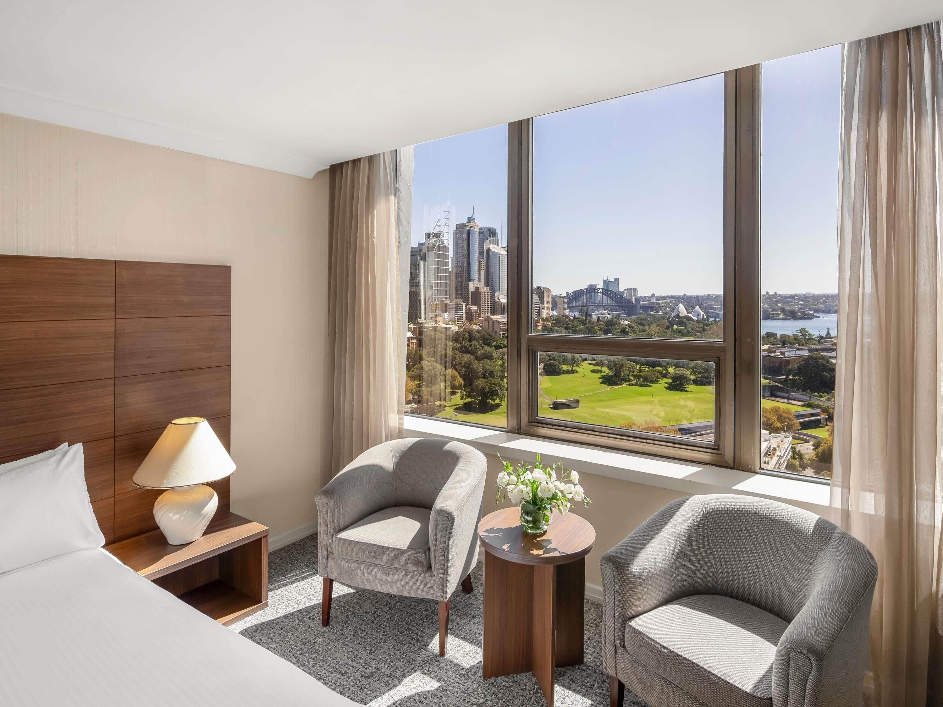 A hotel room with a large window, a bed, two armchairs, a side table, and a lamp.
