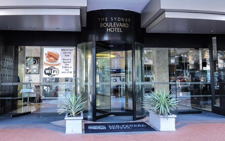 The Sydney Boulevard Hotel entrance with a glass facade, a signboard advertising breakfast, and potted plants.