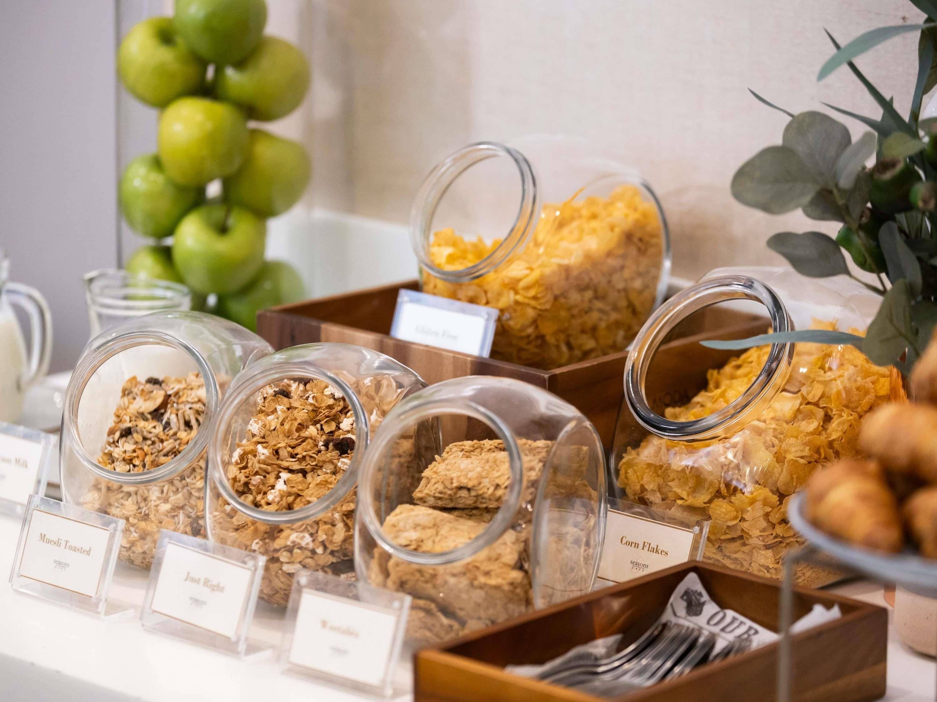 Un bufé con diversos alimentos, entre ellos muesli tostado, copos de maíz y otros cereales, presentados en recipientes transparentes.