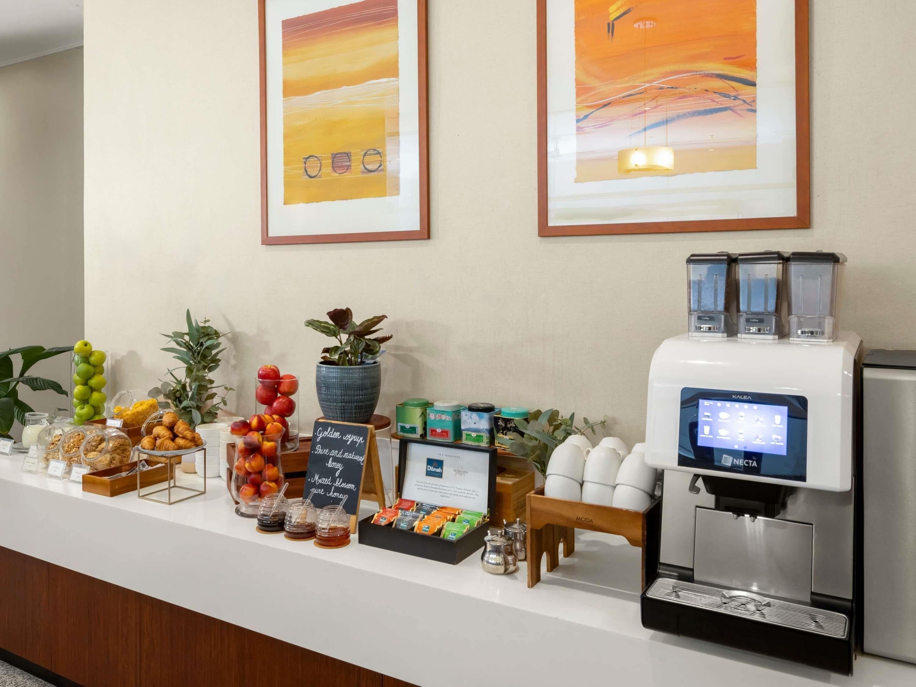 A breakfast buffet with various food items, a coffee machine, and two framed abstract paintings on the wall.
