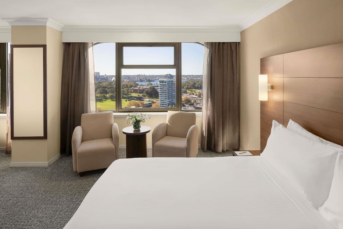 Camera d'hotel con un letto grande, due poltrone, un tavolino con un vaso e vista sulla città attraverso un'ampia finestra.
