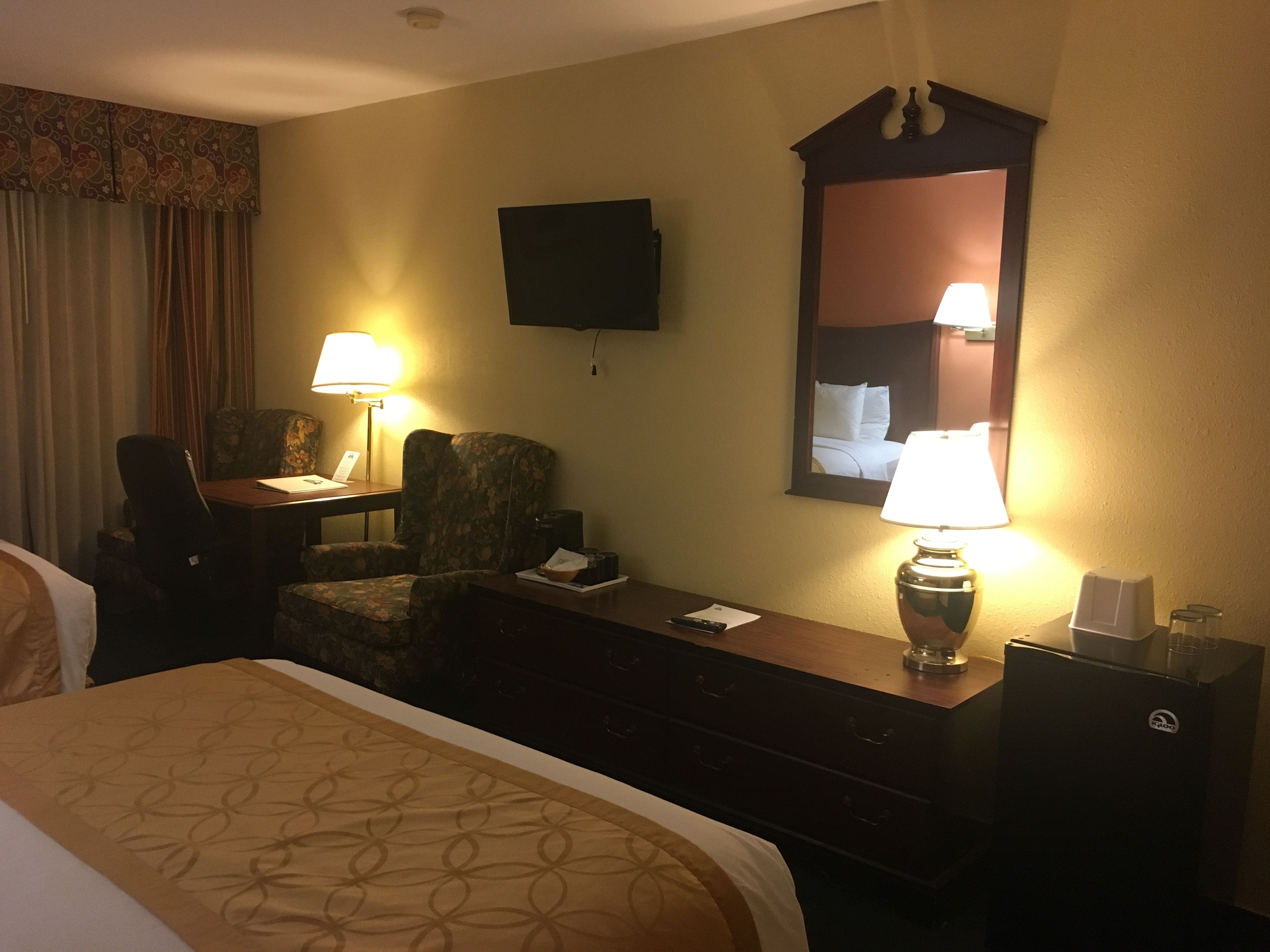 Habitación de hotel con cama, escritorio con silla, televisor, espejo y mesita de noche con lámpara.