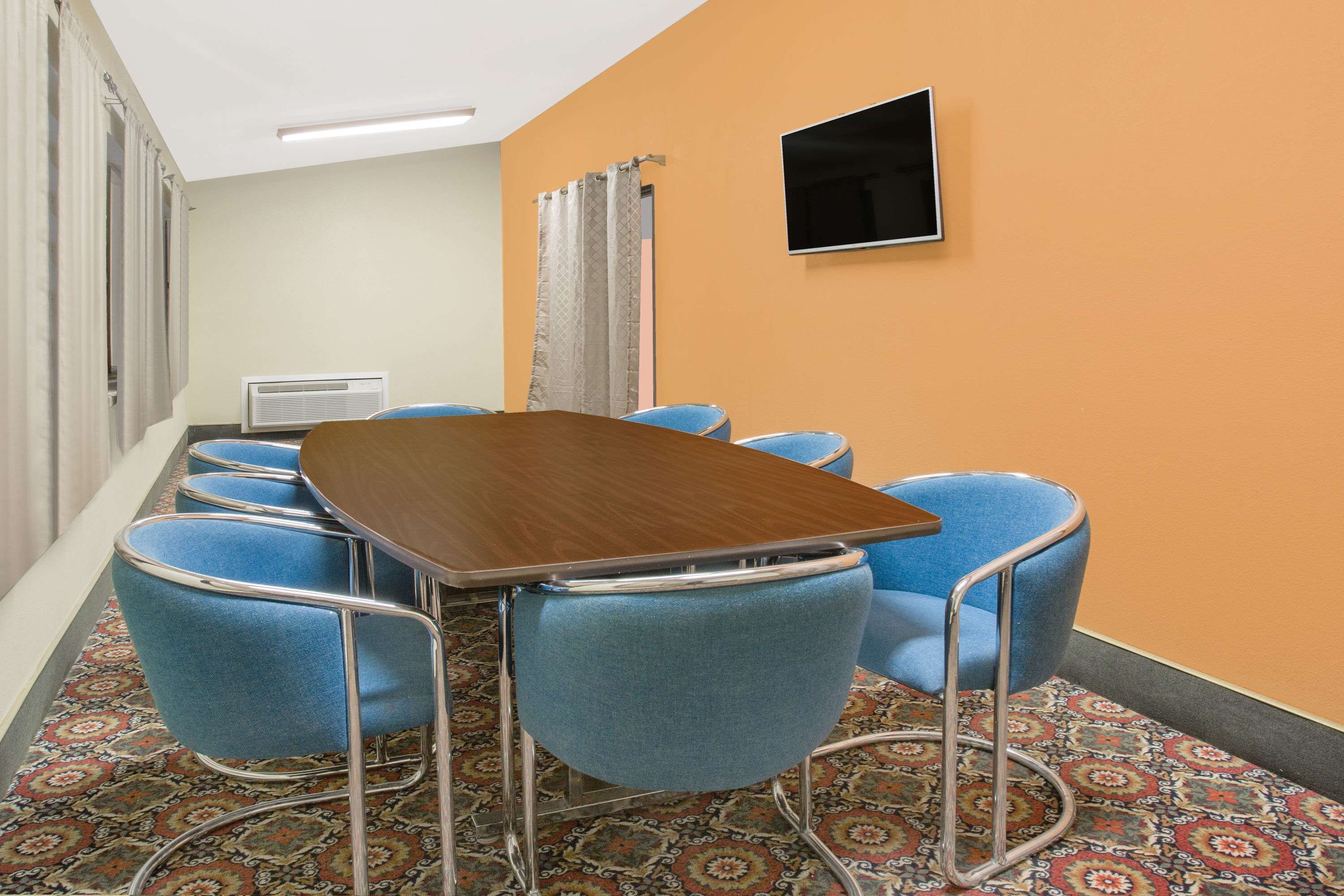A conference room with a wooden table and blue chairs, a flat-screen TV on the wall, and patterned carpet.