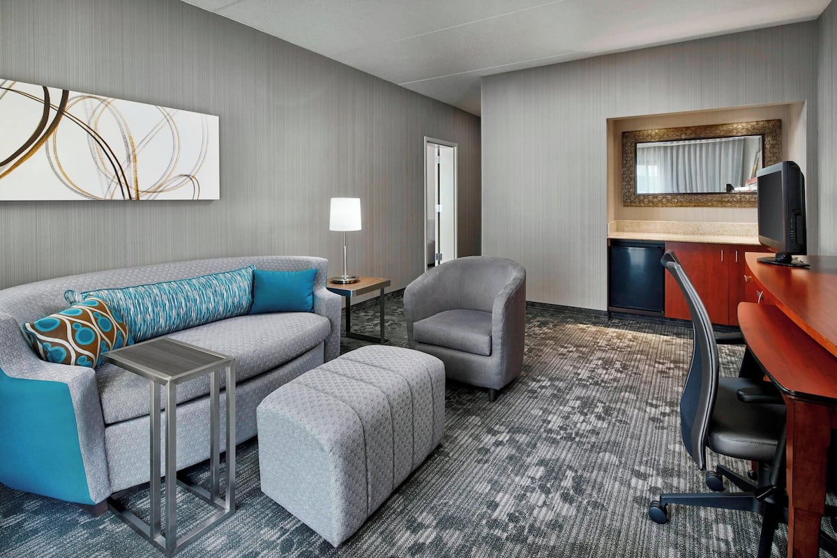 A hotel room with a grey sofa, a grey armchair, a small table with a lamp, a desk with a computer, and a kitchenette area.