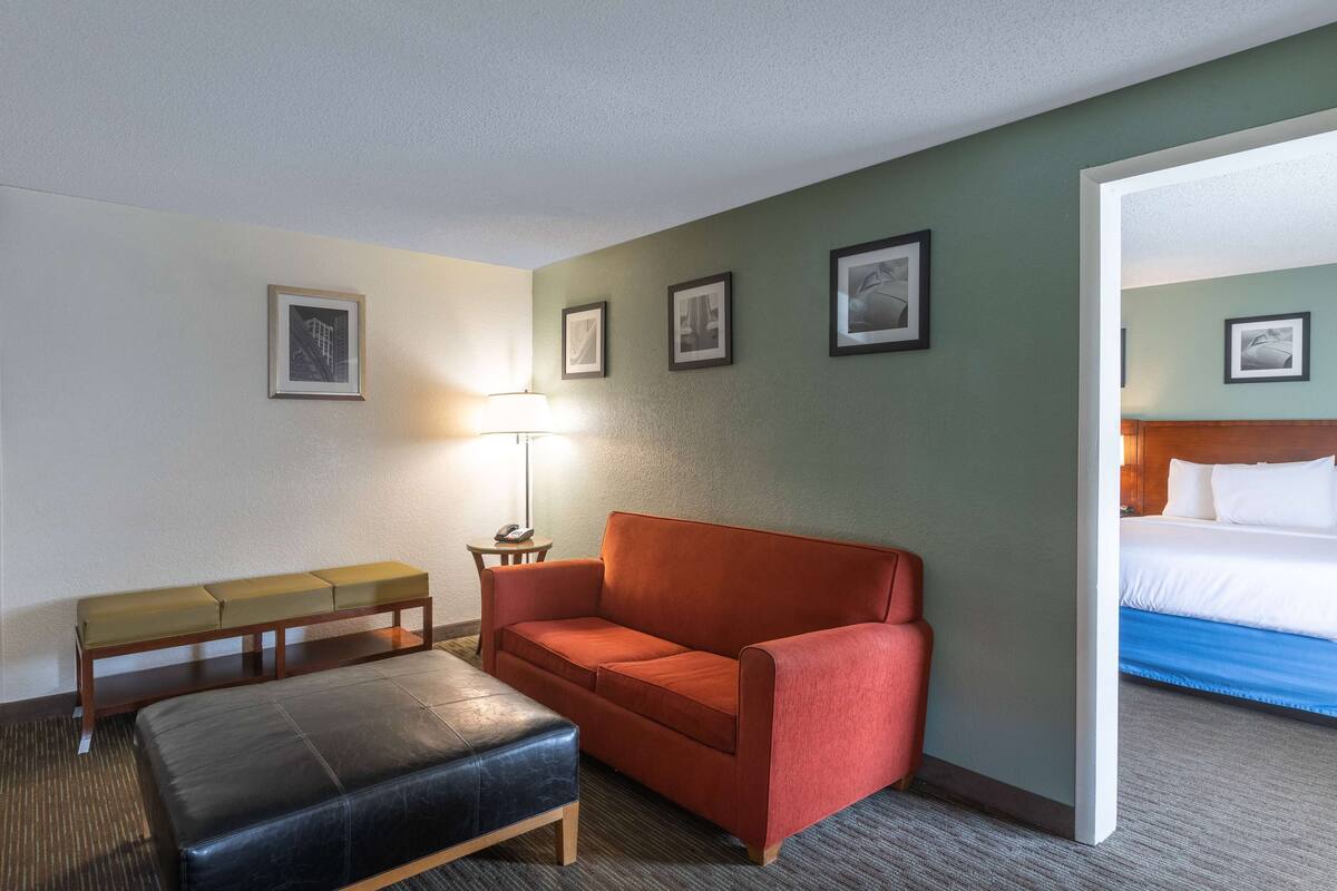 A hotel room with a red sofa, a black leather ottoman, a wooden bench, and a lamp. There are framed pictures on the wall and a bed with white bedding.