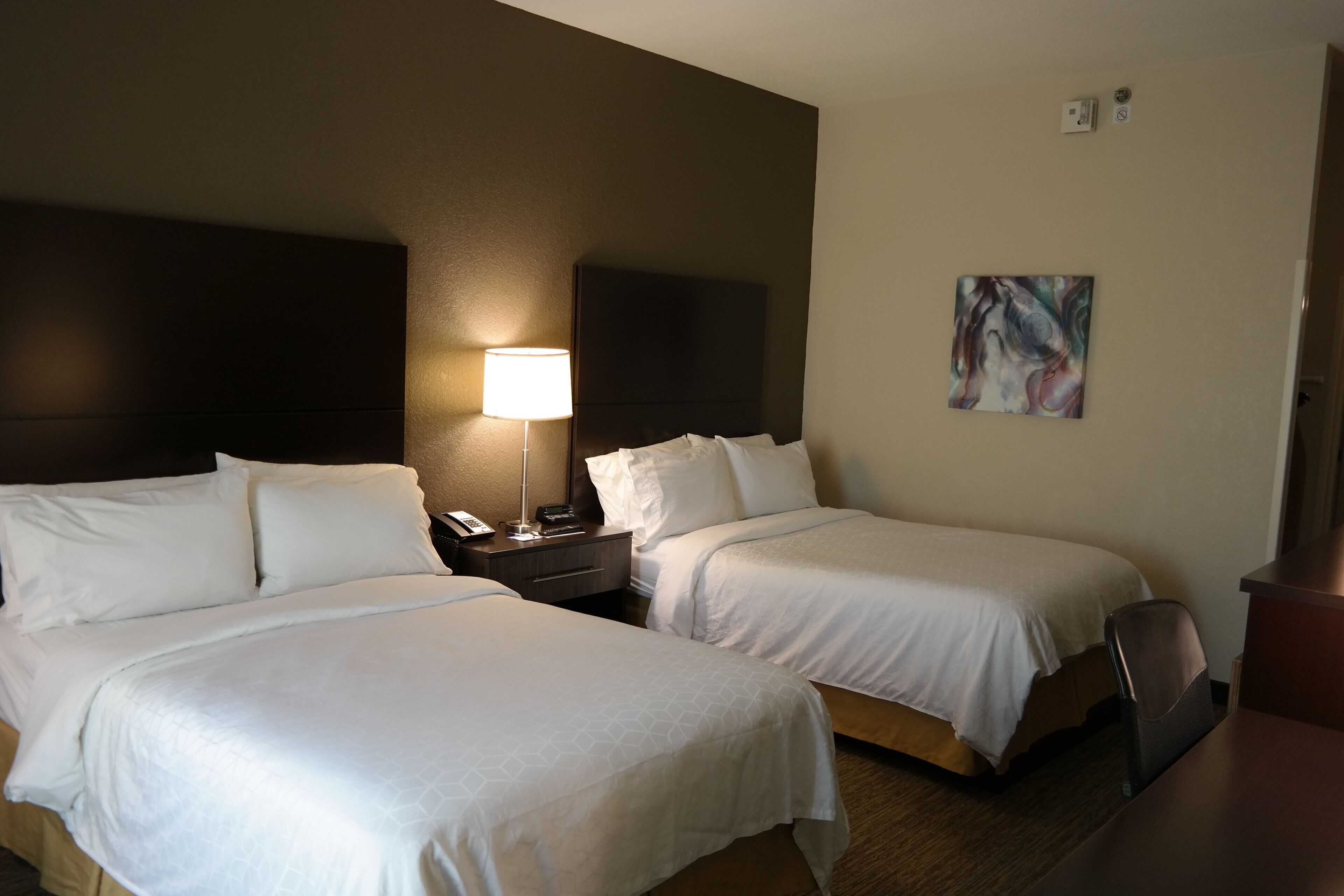 A hotel room with two beds, a nightstand with a lamp, a painting on the wall, and a desk with a chair.