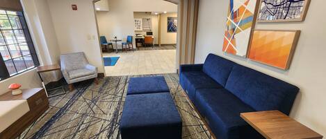 A hotel lobby with a blue sofa, a grey armchair, and a small wooden table. There is a large window on the left, and a door leading to another room.