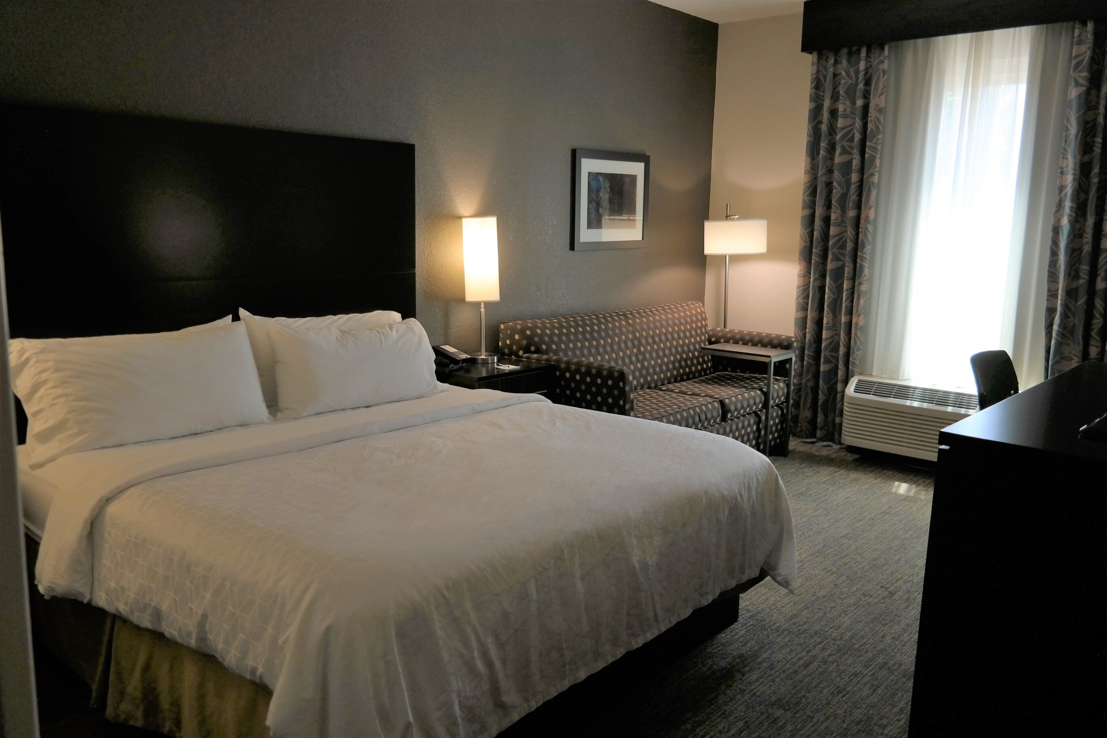 A hotel room with a large bed, a desk, a chair, a standing lamp, and a framed picture on the wall.