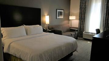 A hotel room with a large bed, a desk, a chair, a standing lamp, and a framed picture on the wall.
