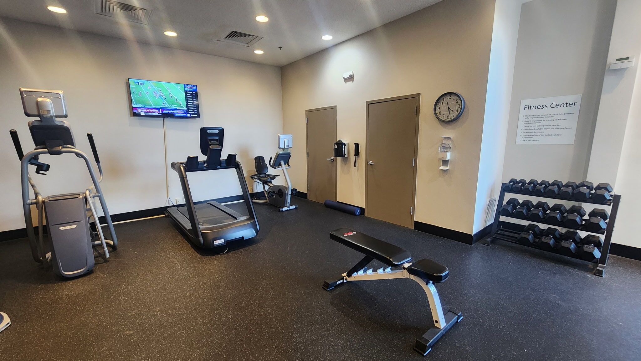 A fitness center with cardio machines, dumbbells, and a TV displaying a sports game.