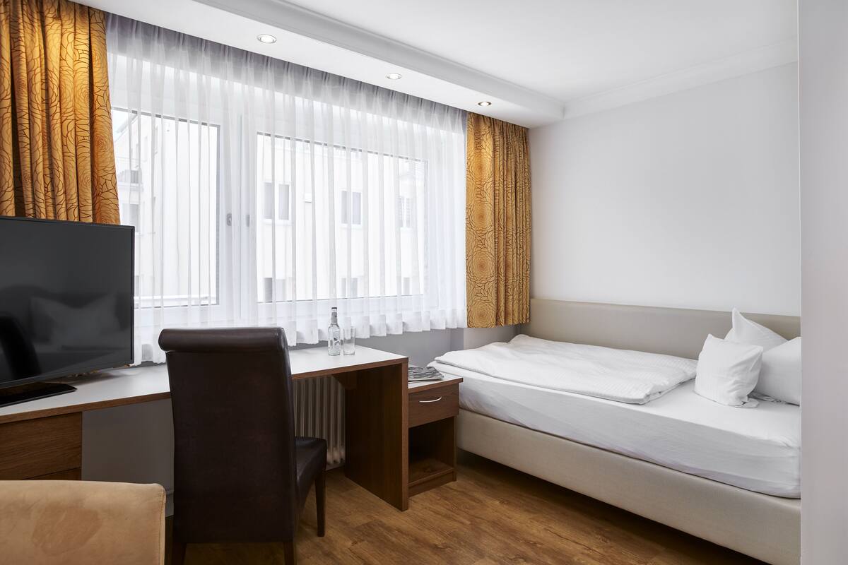 A hotel room with a bed, a desk with a TV, a chair, and a window with curtains.