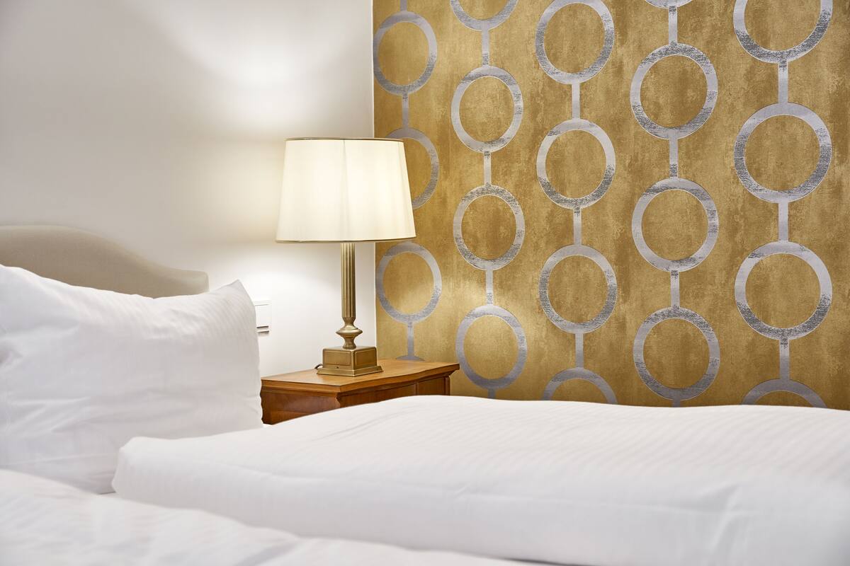 A neatly made bed with white linens, a bedside lamp on a wooden nightstand, and a wall with a geometric pattern.
