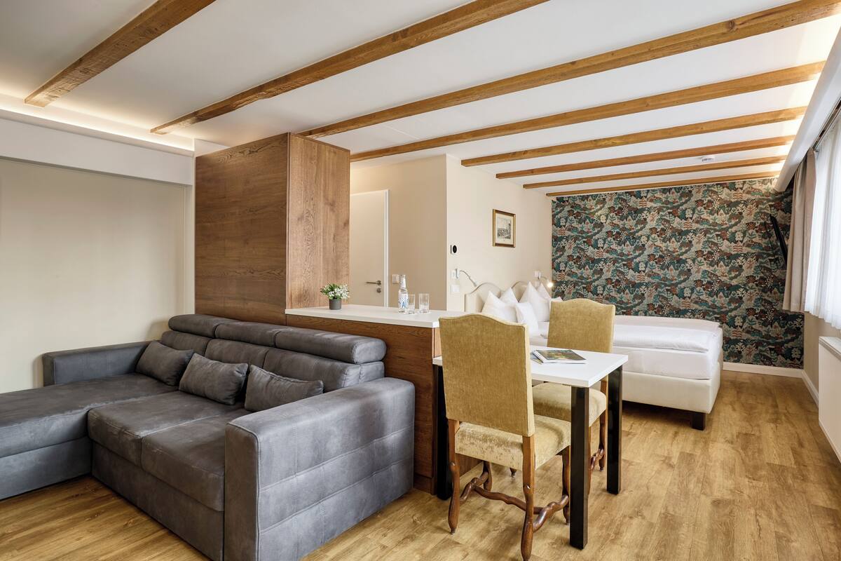 A modern living room with a sectional sofa, a dining table with chairs, wooden beams on the ceiling, and a patterned wallpaper wall.