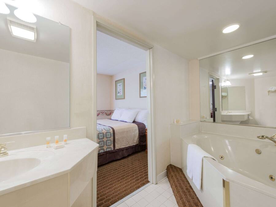 Un baño moderno con una bañera grande, un lavamanos y un espejo. Se ve una puerta que da a un dormitorio con cama y piso alfombrado.