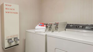 A laundry area with a coin-operated soap, bleach, and softeners dispenser, a stack of white towels, and a front-loading washing machine. - Days Inn by Wyndham Yadkinville (Yadkinville)