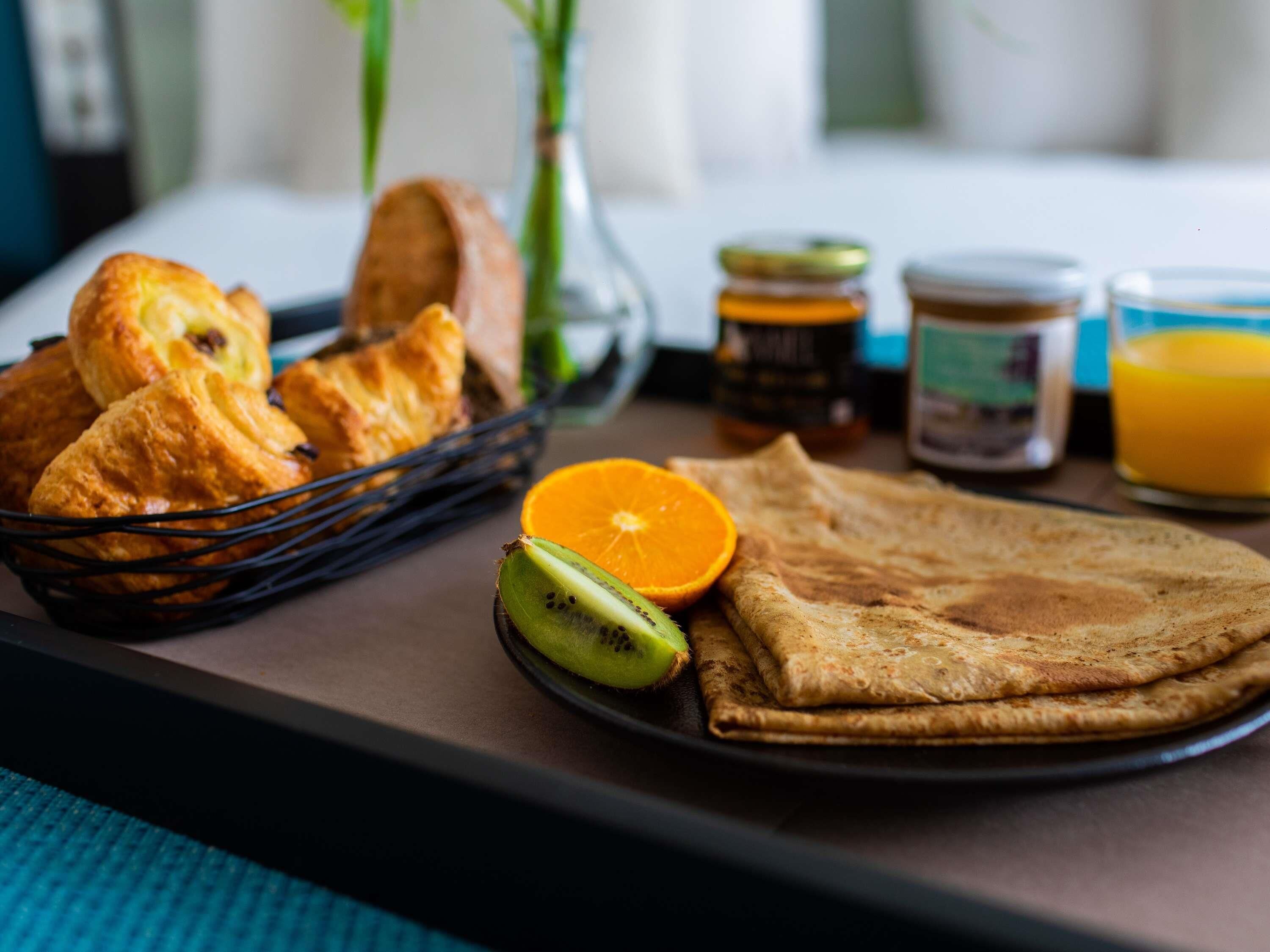 Bandeja de desayuno con cruasanes, una crepa enrollada, un kiwi, una rodaja de naranja y un vaso de jugo de naranja.