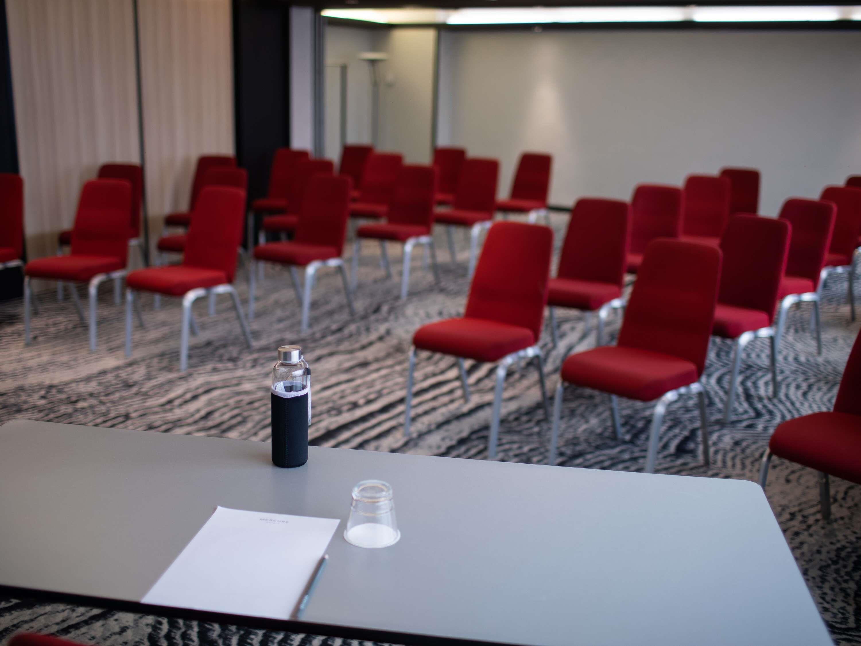 A conference room with rows of red chairs, a table with a water bottle and a glass, and a whiteboard in the background.