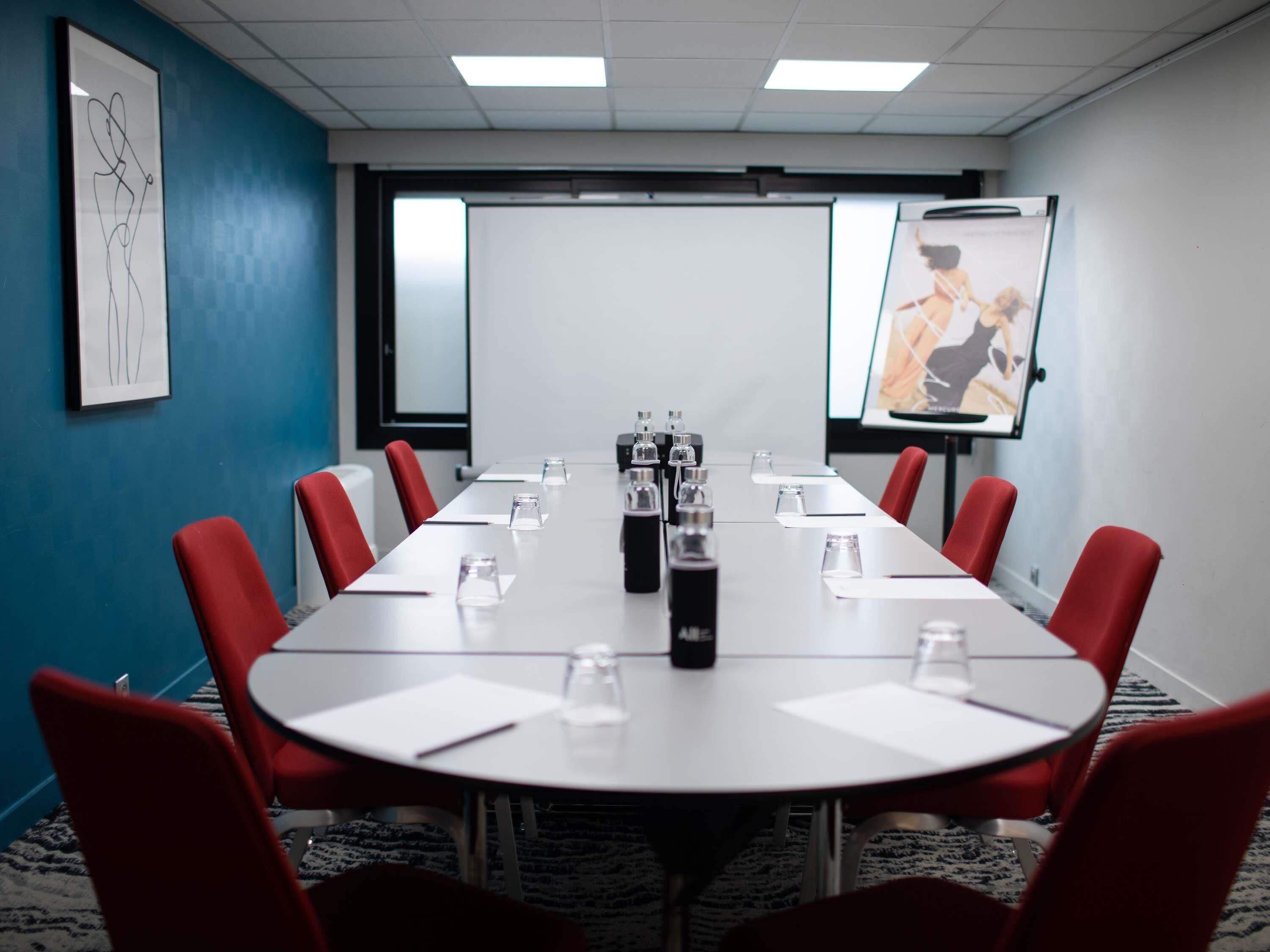 A conference room with a long table, red chairs, water bottles, and a projector screen.