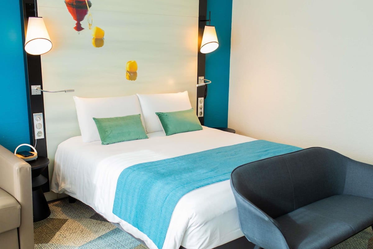 A neatly arranged hotel room with a bed, a chair, a nightstand, and a wall-mounted lamp.