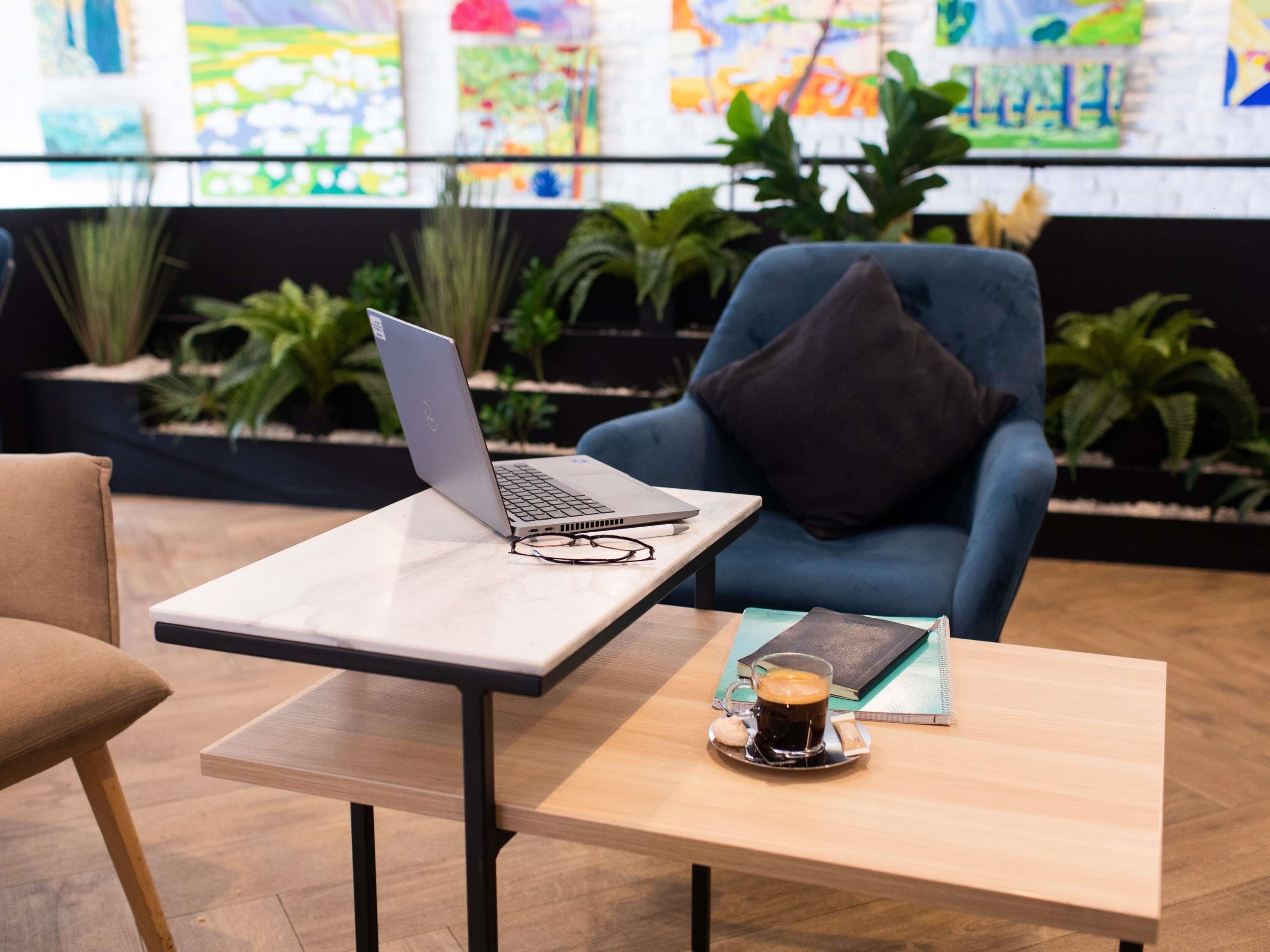 A modern indoor space with a laptop on a table, a chair with a cushion, a coffee cup on a small table, and potted plants in the background.