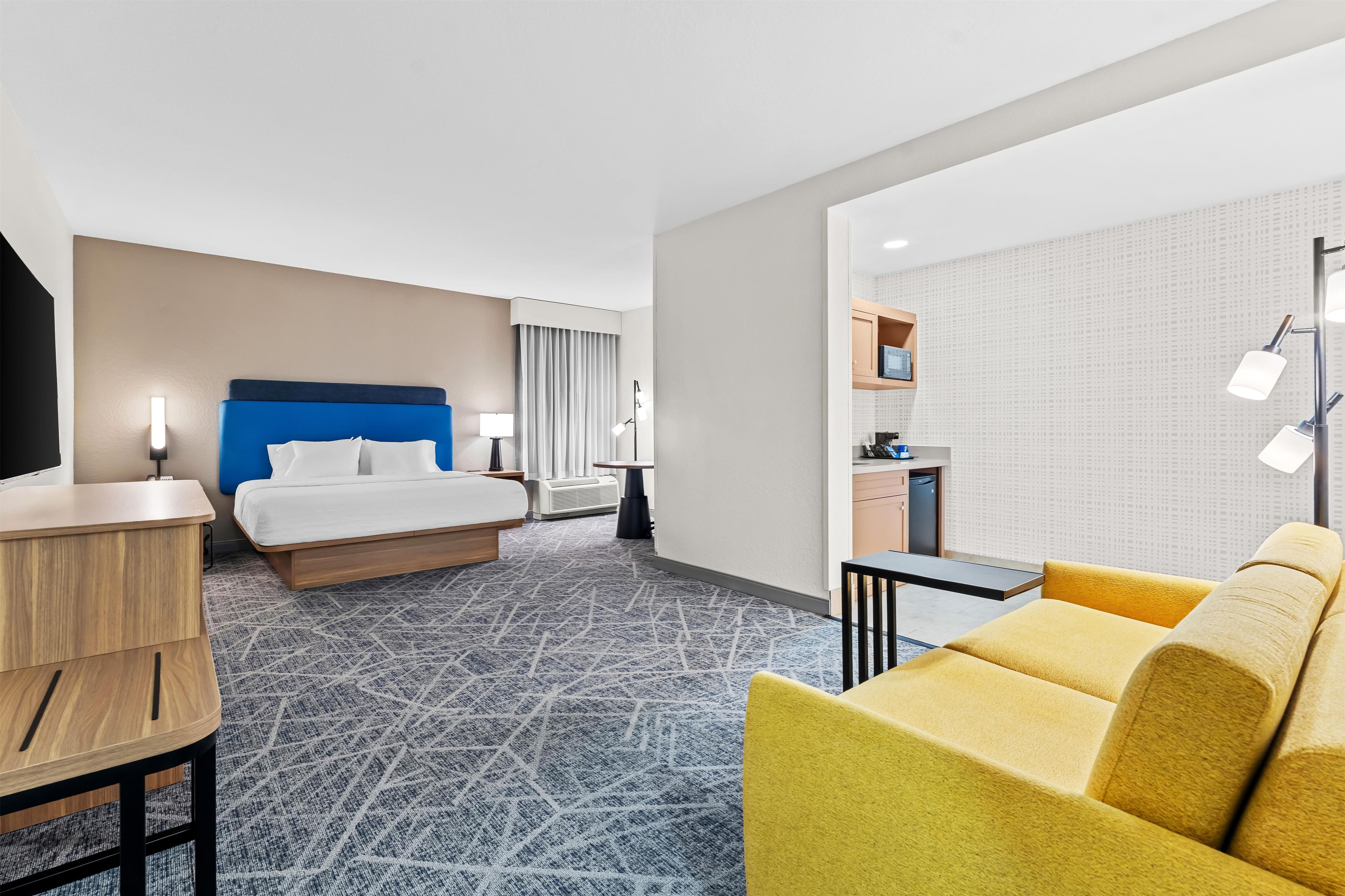 A hotel room with a bed, a desk, a sofa, a kitchenette, and a patterned carpet.