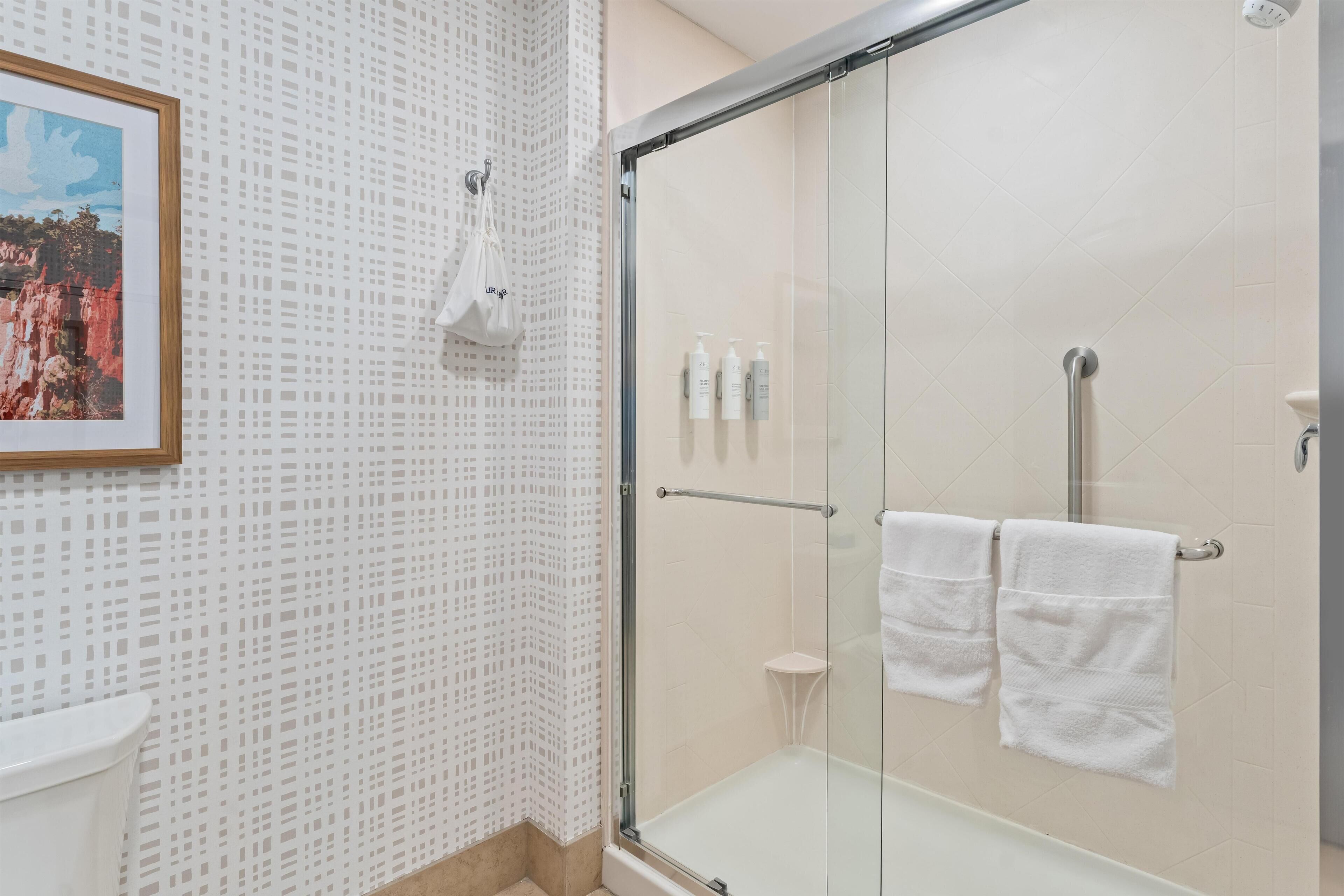Uma casa de banho moderna com cabine de duche em vidro, um quadro emoldurado na parede e um banquinho branco pequeno.