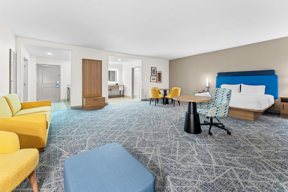 Ein Hotelzimmer mit einem Bett, einem Schreibtisch, zwei gelben Stühlen, einem blauen Hocker und einem gemusterten Teppich.
