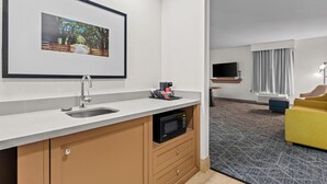 A modern hotel room with a kitchenette, a flat-screen TV, a yellow armchair, and a large window with curtains. - Hampton Inn & Suites Tifton (Tifton)
