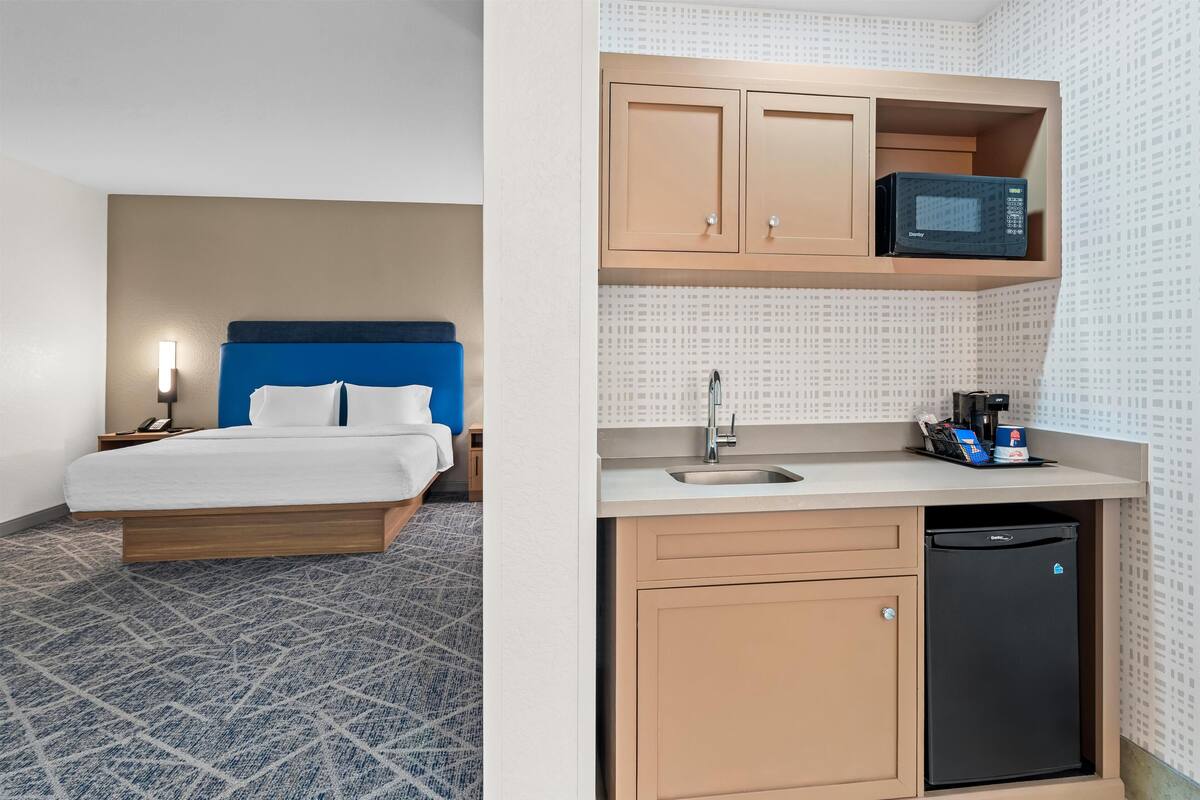 Ein Hotelzimmer mit einem Bett, einem Nachttisch, einer Wandlampe und einer Küchenzeile mit Mikrowelle, Spüle und Kühlschrank.