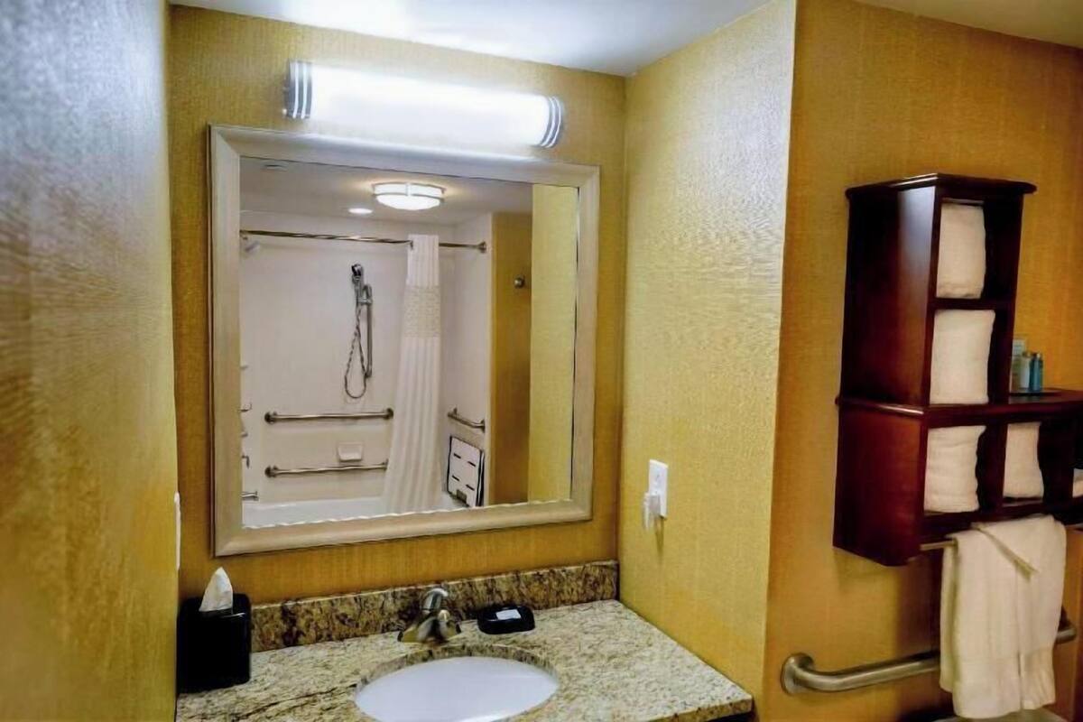 Ein Badezimmer mit Waschbecken, Spiegel, Handtuchhalter und einer in der Spiegelung sichtbaren Dusche.