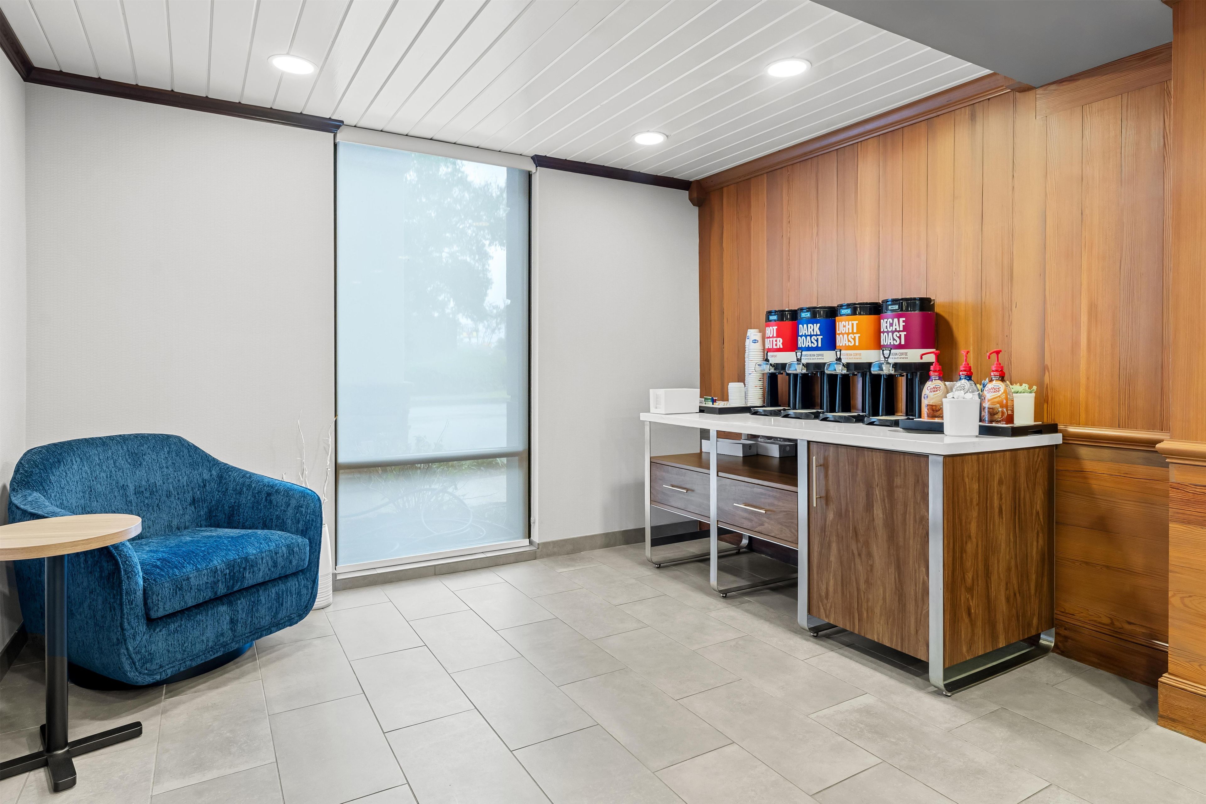 A seating area with a blue chair and a small table, a coffee station with dispensers, and a view through a glass door.