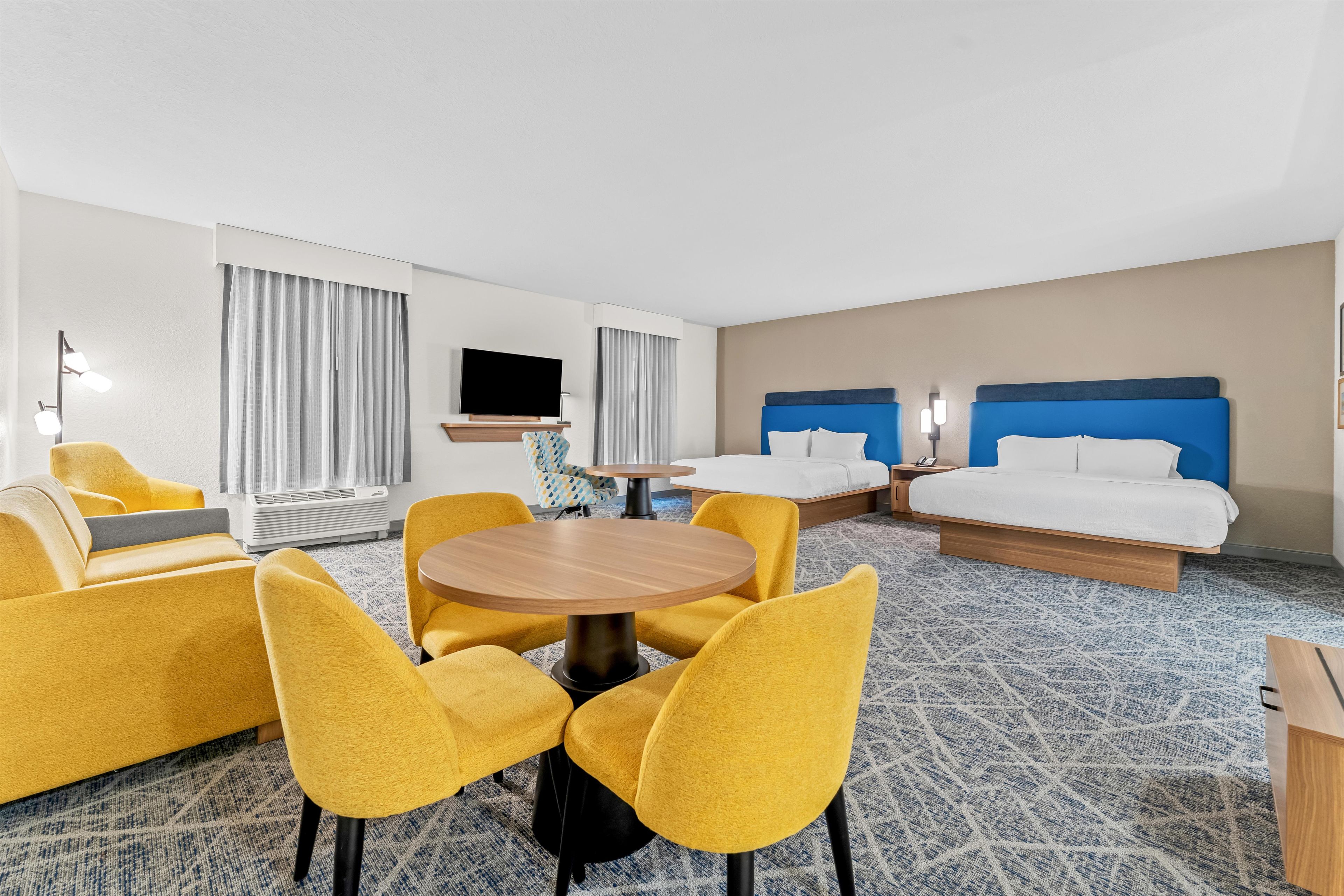 A hotel room with a bed, two yellow armchairs, a round table, a TV, and a window with curtains.