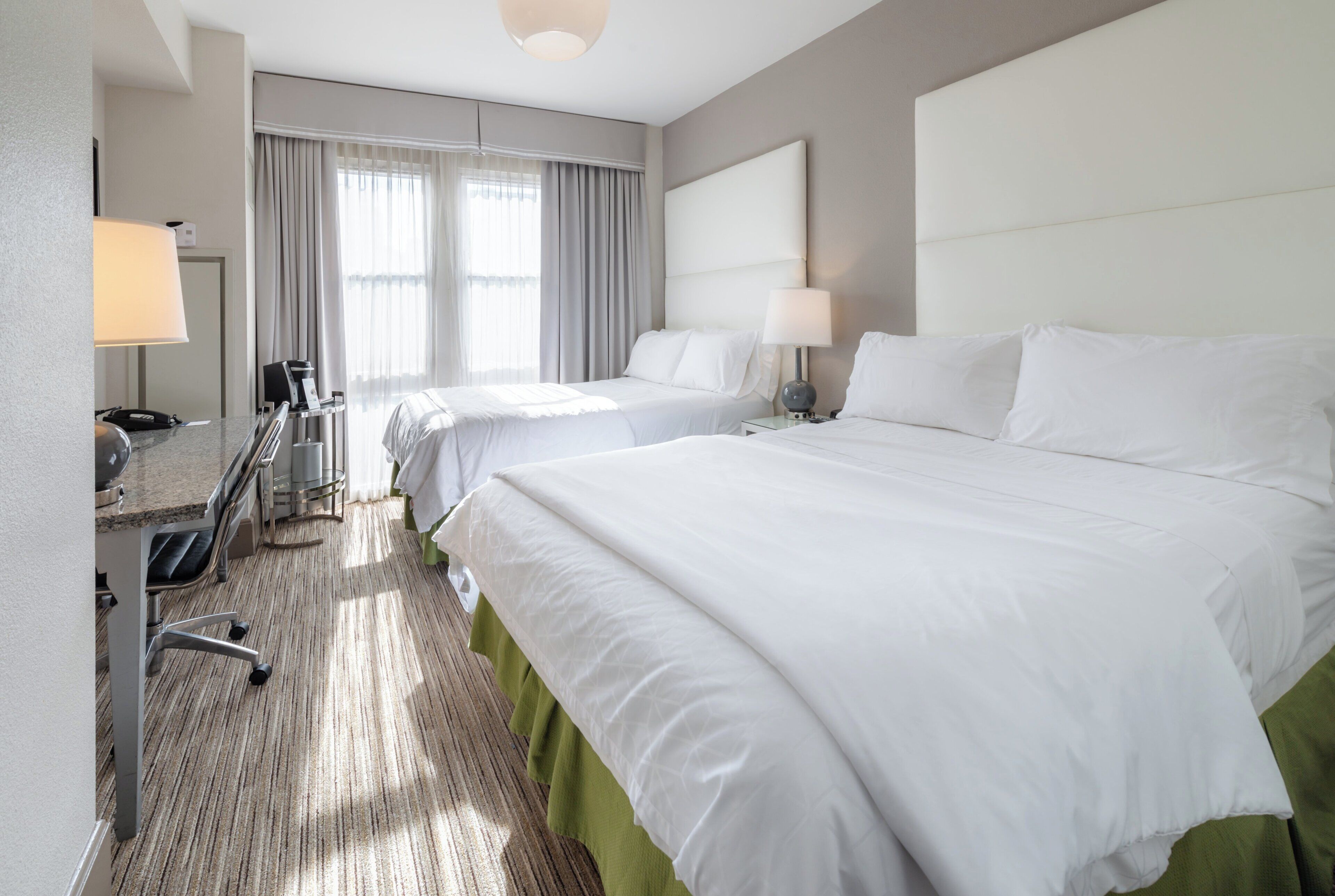 Habitación de hotel con dos camas, un escritorio con lámpara, una silla, cafetera y ventana con cortinas.