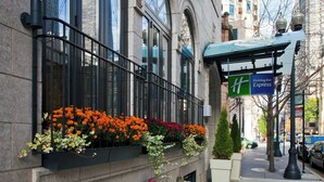 Un bâtiment doté d’un balcon orné de jardinières remplies de différentes sortes de fleurs, une enseigne indiquant un hôtel Holiday Inn Express, et une vue de la rue avec des voitures garées et des bâtiments en arrière-plan.
