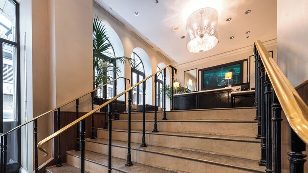 Un magnifique escalier en marbre, avec des marches en marbre et des balustrades noires, mène à un hall d’entrée agrémenté d’un lustre et de grandes fenêtres.