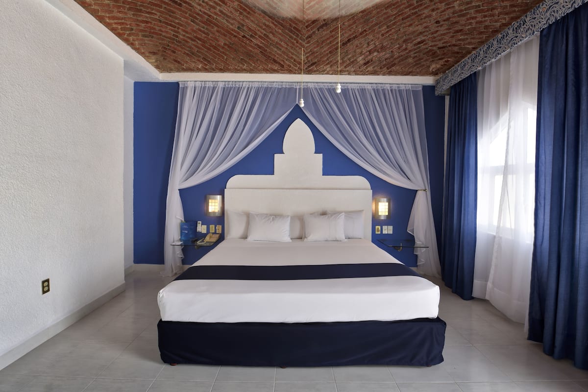 A large bed with a white headboard and blue curtains in a room with blue walls and a brick ceiling.