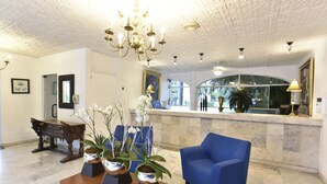 Un espace d’accueil avec un fauteuil bleu, une table en bois ornée de pots d’orchidées, un lustre et un comptoir de réception en marbre.