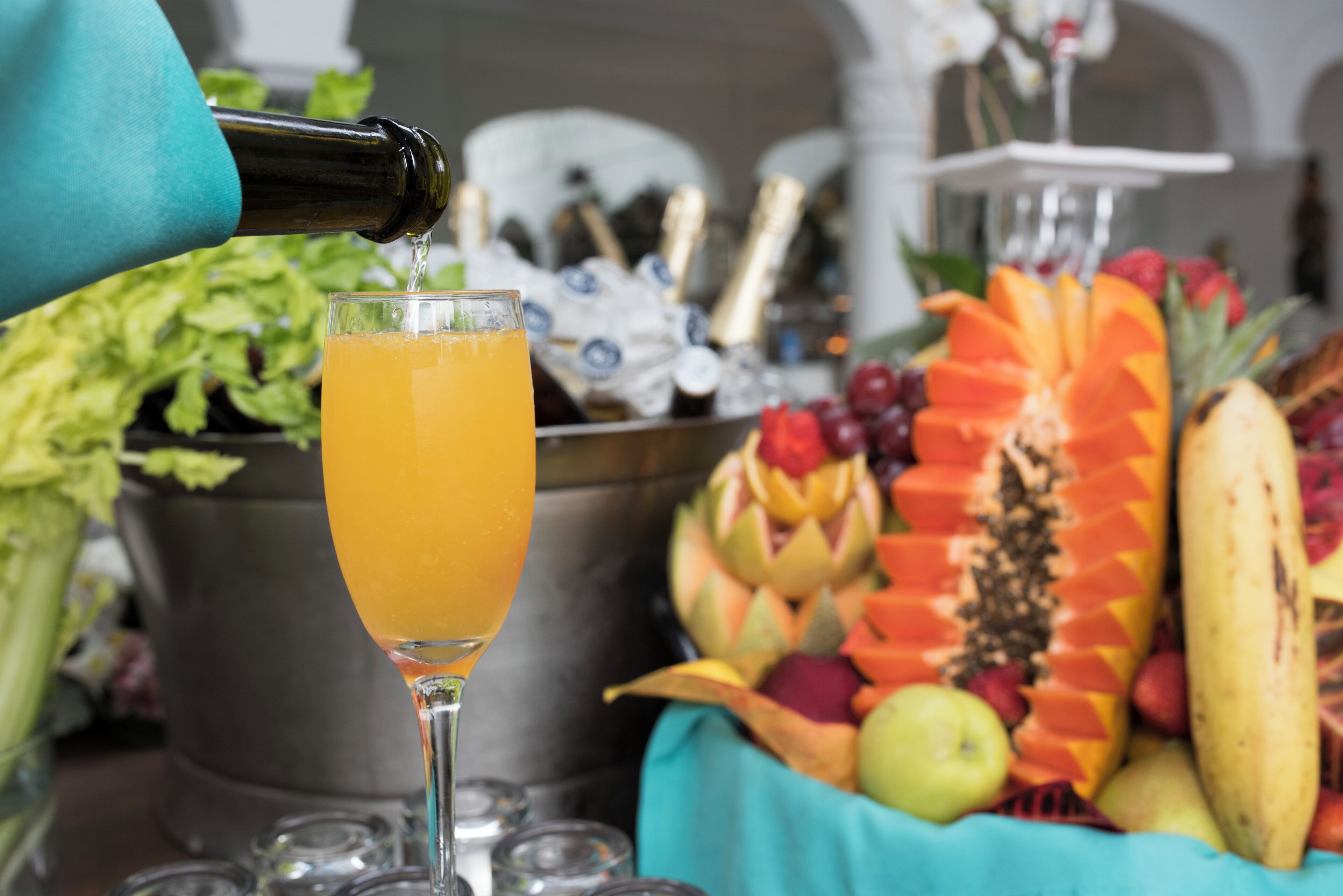 Una copa de cóctel siendo servida con jugo de naranja, rodeada de una exhibición de frutas que incluye una piña tallada y un plátano.